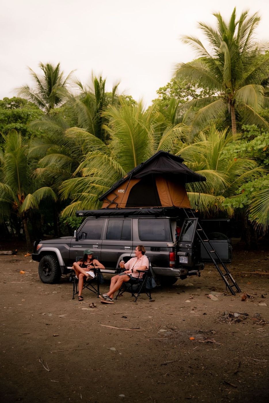 Couple sitting in camp chairs beside a Toyota Land Cruiser LX with its rooftop tent open