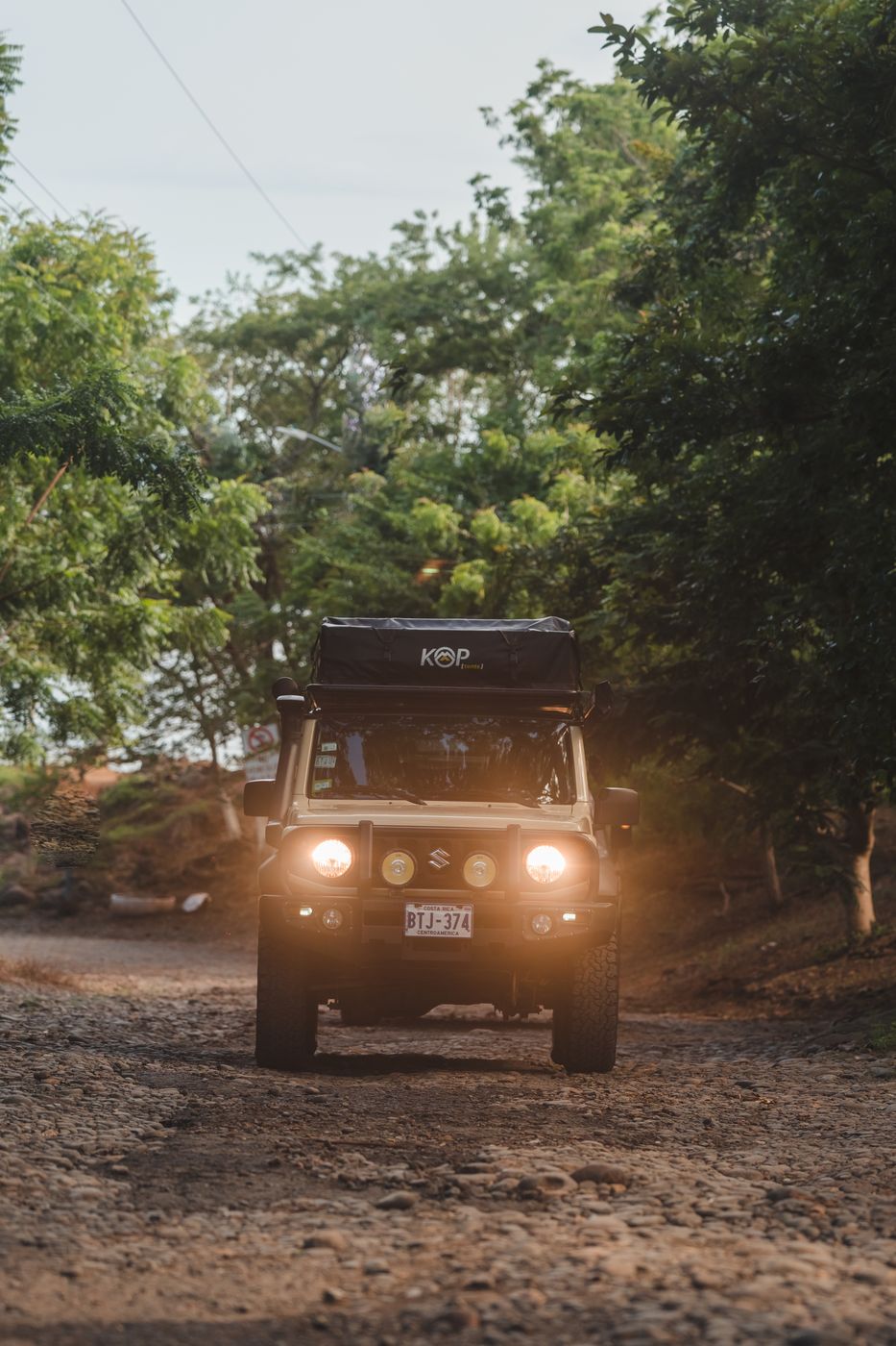 Suzuki Jimny with its lights on, driving a dirt road