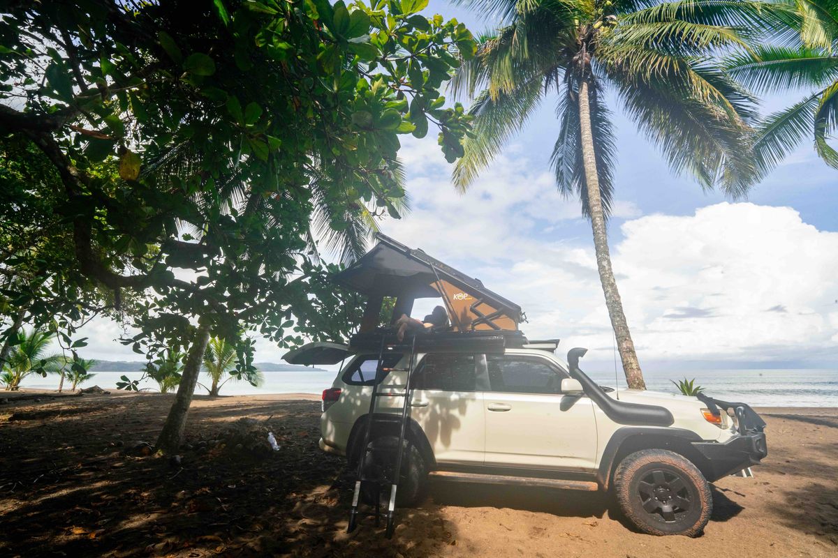 White 4-door Toyota 4x4 with a rooftop tent parked under palms on a beach